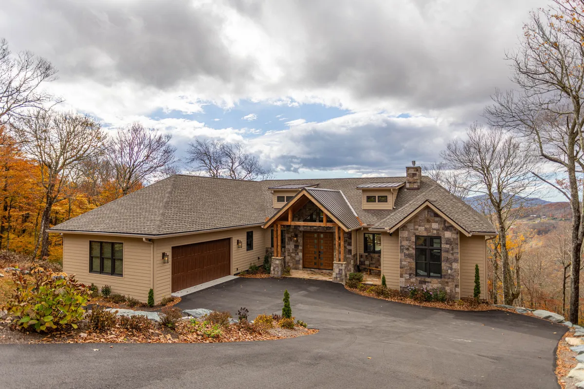 The image showcases the grand front entrance of a luxurious single-family home set on Sugar Mountain, featuring elegant stone accents, a striking wooden double door, and beautifully landscaped gardens with mountain vistas visible in the background. The setting glows with sunset light, hinting at spectacular evening views and inviting outdoor spaces.