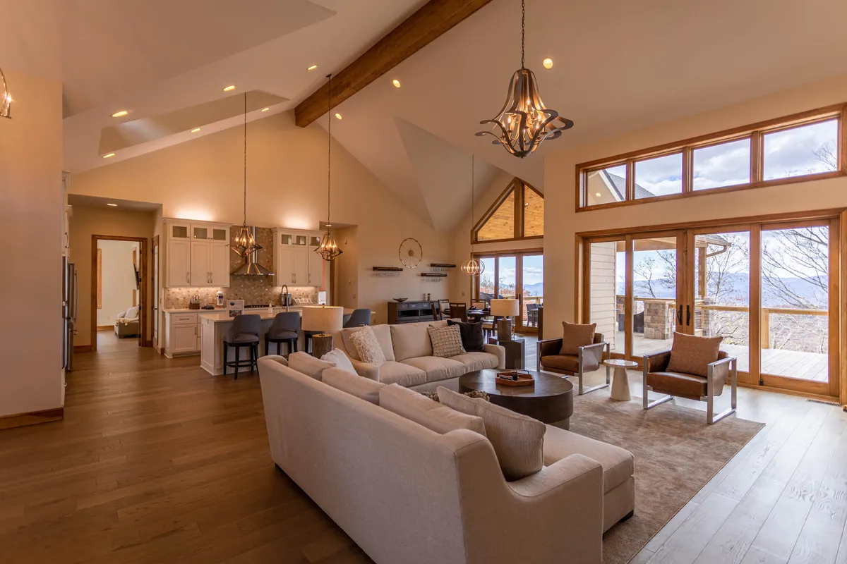 A spacious great room with a large sectional sofa positioned beneath a grand chandelier and striking angled windows. The wooden beams accentuate the cathedral ceiling, while the panoramic windows showcase lush scenery outside. The layout encourages gathering and togetherness.