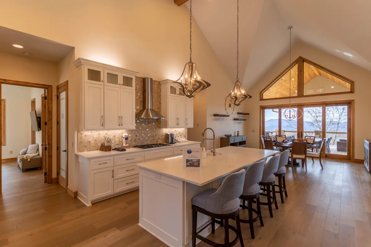 The scene focuses on the kitchen island with a view extending into the sunlit dining area. Large windows offer glimpses of the outdoors, and the layout suggests a seamless flow between cooking, dining, and enjoying the surrounding landscape. The mood is cozy, sophisticated, and perfectly suited to festive meals or casual brunches.
