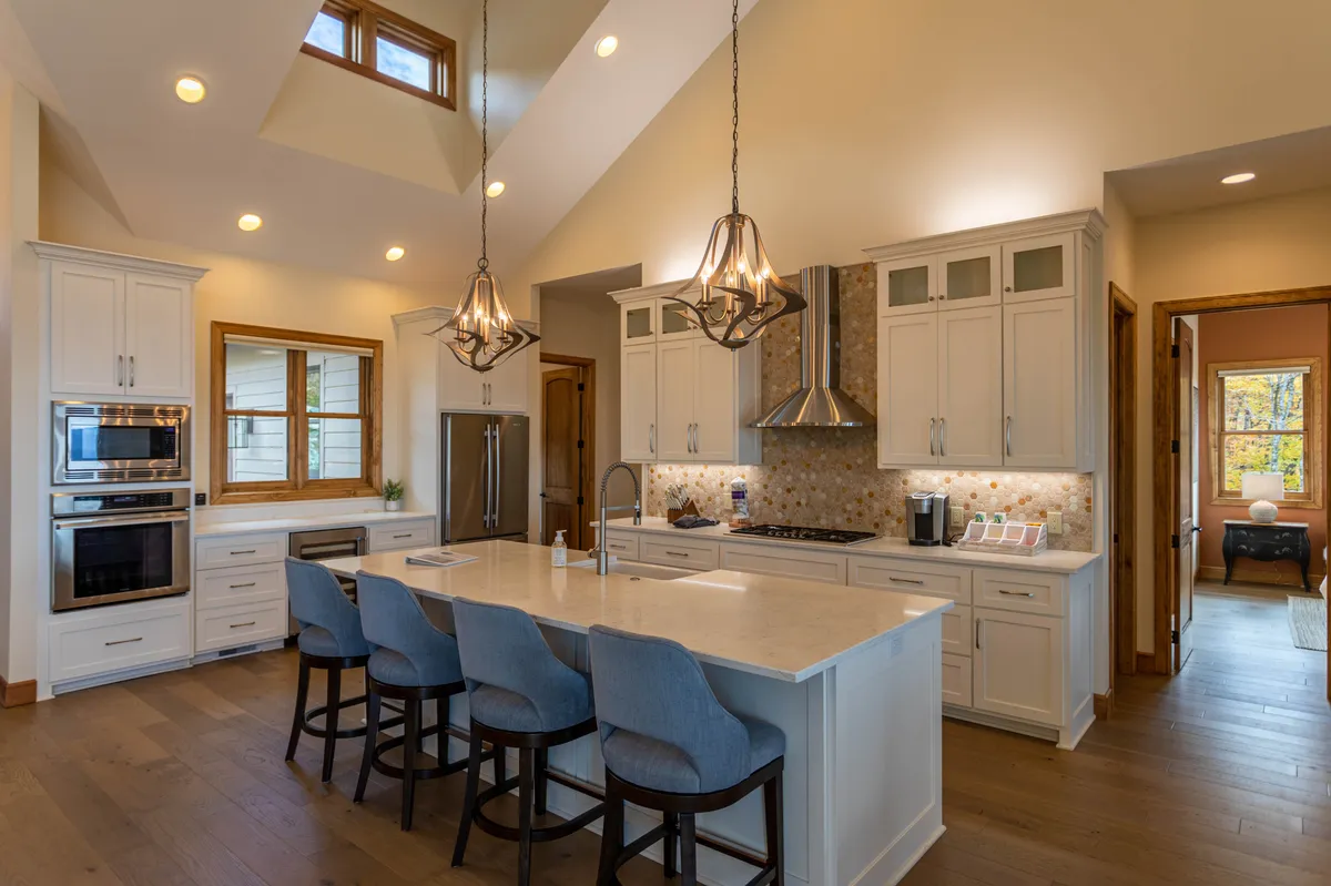 This image showcases a contemporary kitchen with a spacious central granite island, modern appliances, elegant light fixtures, and natural light streaming in from nearby windows. The island has ample seating, and the setting exudes a welcoming, functional atmosphere ideal for cooking and entertaining.