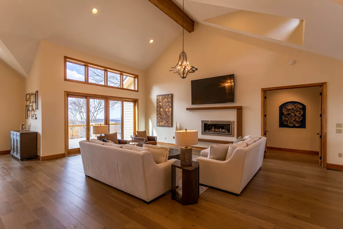 This image features a plush sofa seating arrangement centered around a glass-enclosed gas fireplace and a state-of-the-art LED TV. The area is bathed in natural light from expansive, floor-to-ceiling windows that frame breathtaking mountain views. The overall mood is cozy and welcoming, perfect for relaxation and socializing.