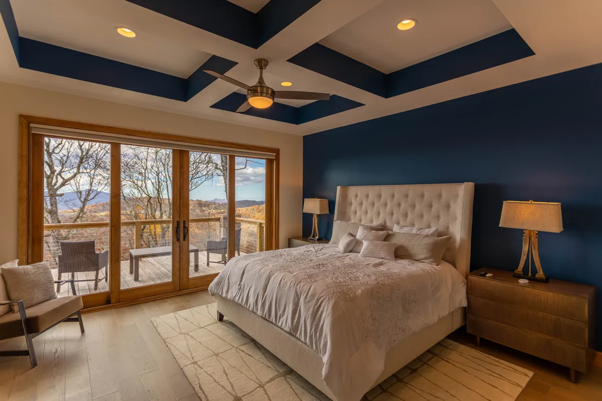 A luxurious bedroom featuring a tufted headboard on the king bed, large sliding glass doors opening to a private deck, decorative ceiling design, and nightstands with soft lighting. Natural light fills the room, highlighting a tranquil atmosphere ideal for relaxation.