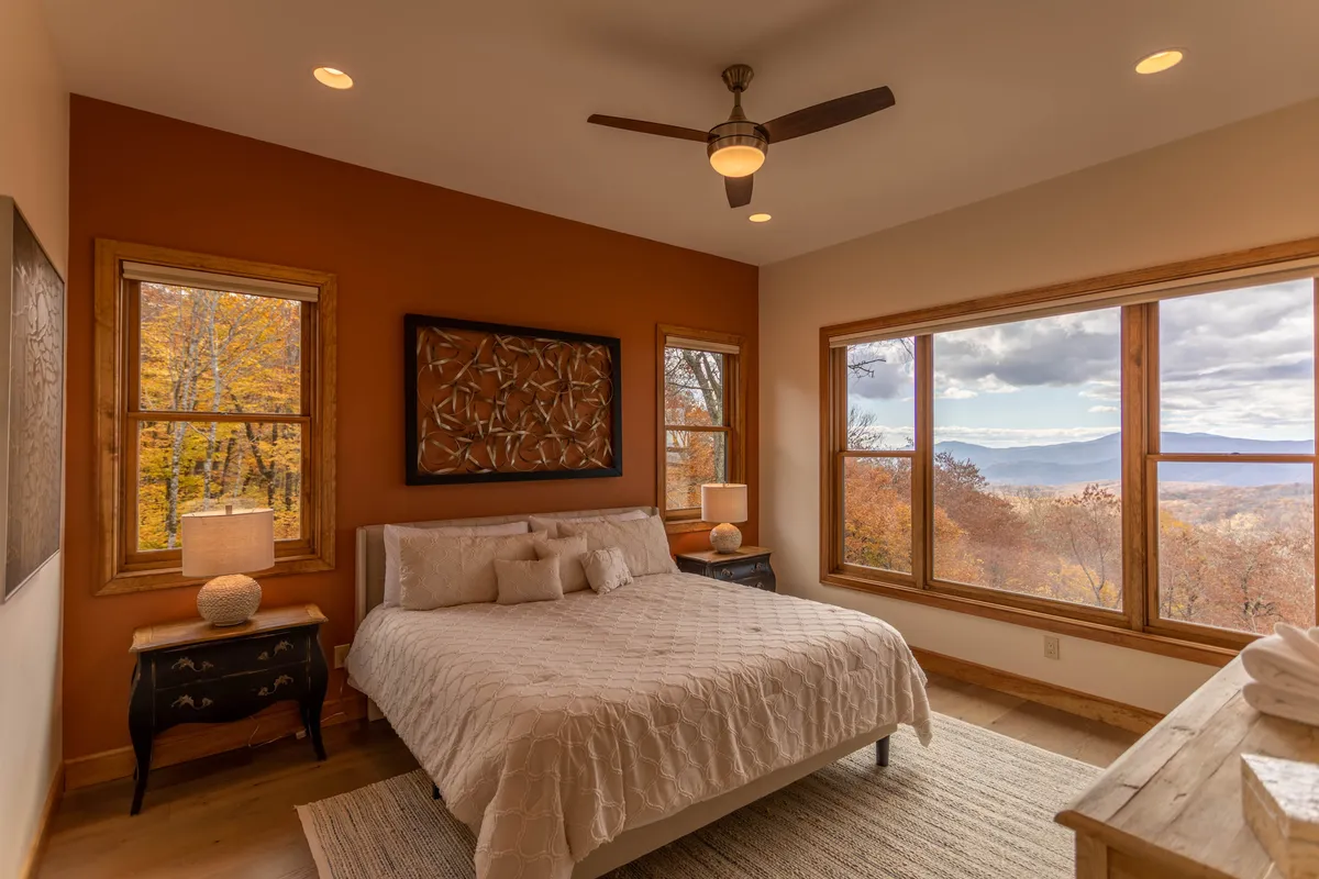 A serene king bedroom with white bedding, large windows framing scenic views, wooden nightstands with lamps, and decorative wall art. The space feels airy, inviting, and perfectly suited for restful evenings or tranquil mornings.