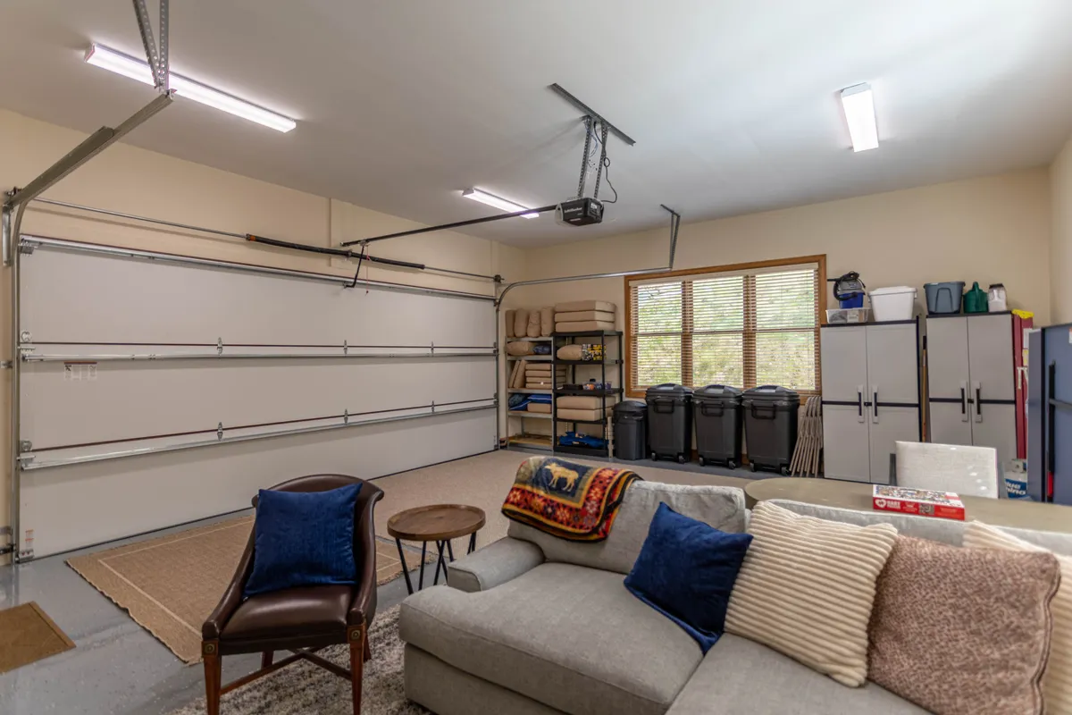 A converted garage space with a visible garage door, a comfortable couch, storage shelves, and a window that brings in natural light. The setup suggests flexibility, with open floor space that could accommodate a ping pong table or other recreational activities, as well as social gatherings.